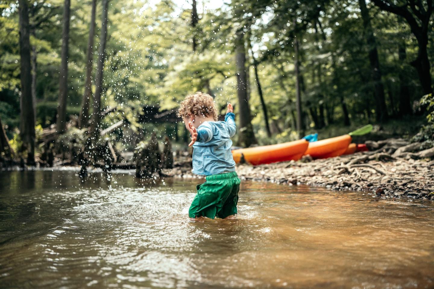 Child splashing in a forest river near orange kayaks.