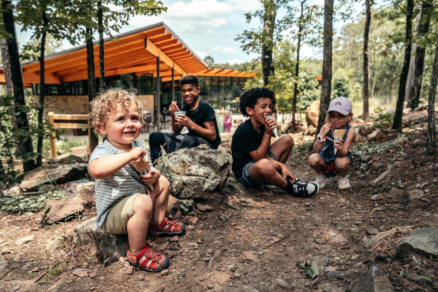 Children sitting on rocks, smiling outdoors near a wooden building and trees.