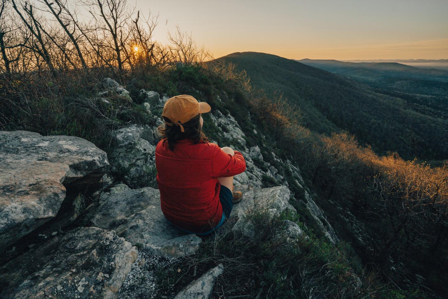 Person in red jacket sitting on rocky hill at sunset, overlooking forested valley.