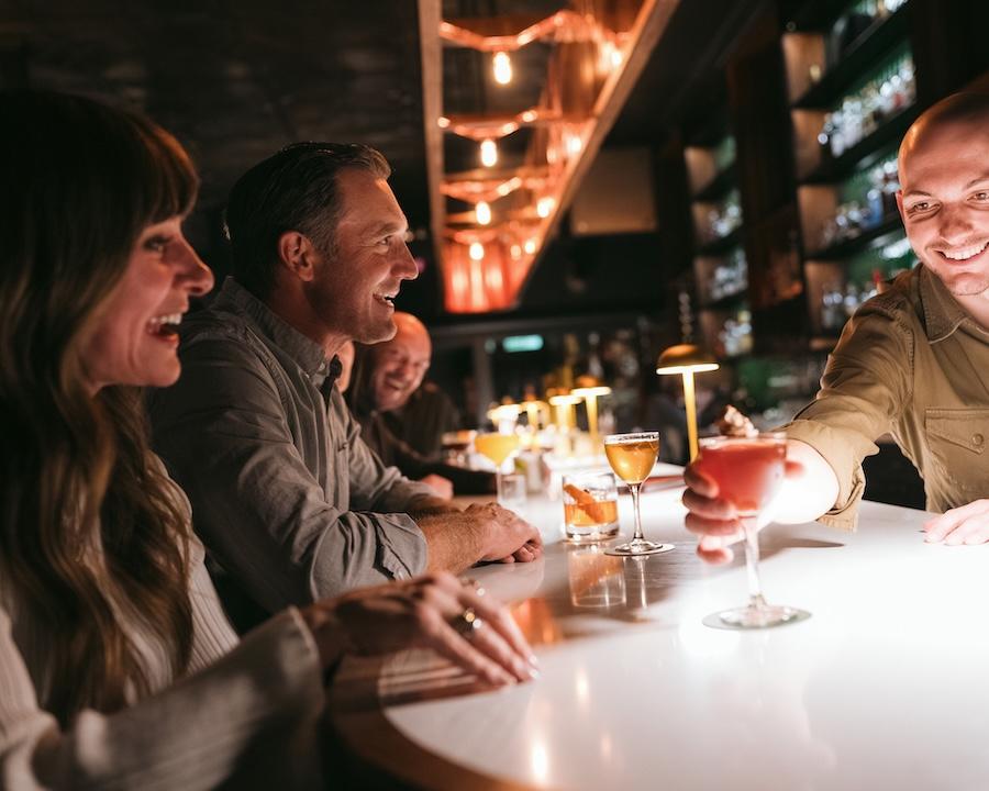 Friends enjoy drinks at a warmly lit bar, smiling and talking.