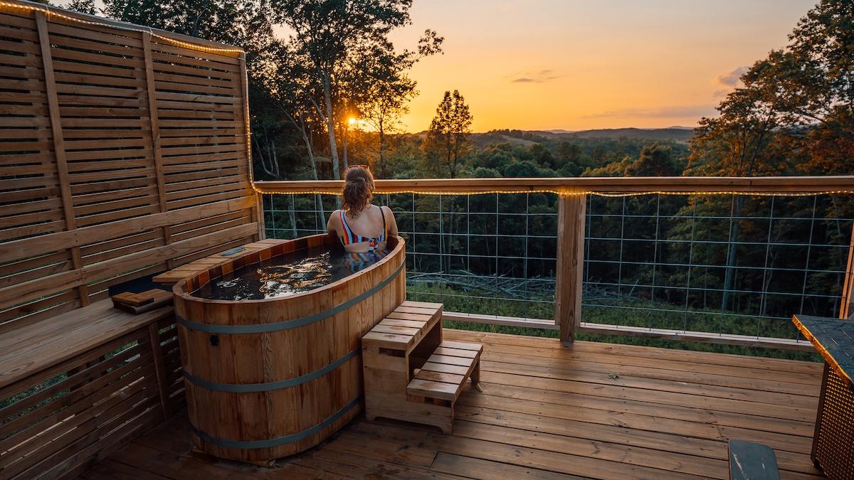 Woman in hot tub on deck, watching sunset over trees.