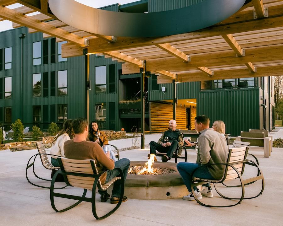 Group sitting around an outdoor fire pit, modern building in the background.