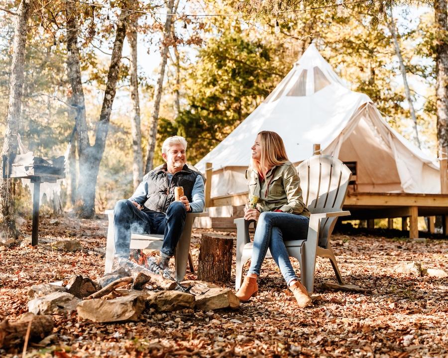 Two people sitting by a campfire near a tent in an autumn forest.