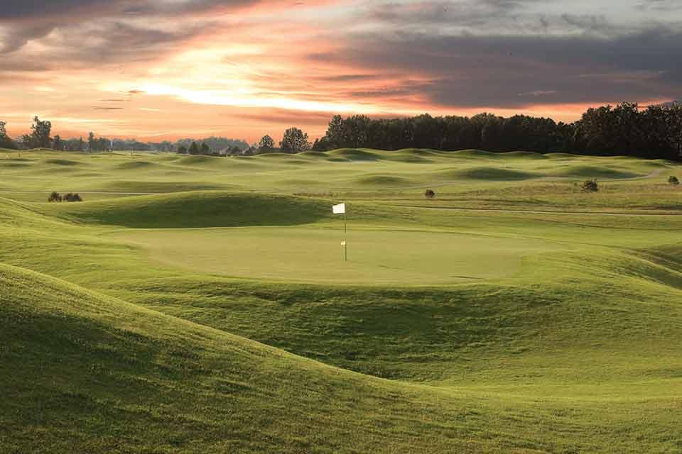 Golf course at sunset with rolling green hills and a distant pin flag.