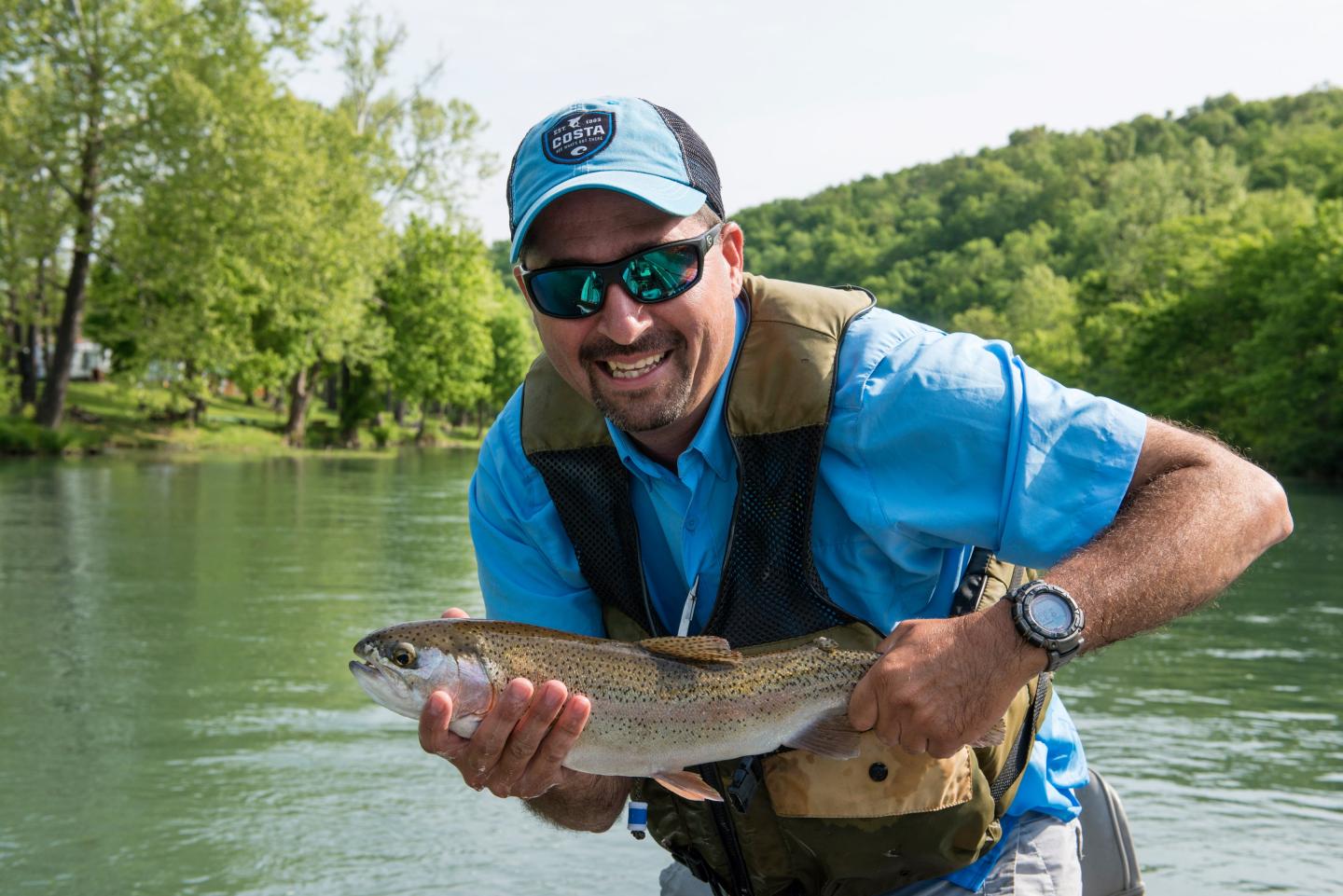 Man smiling with caught fish by a river, wearing sunglasses and a cap.