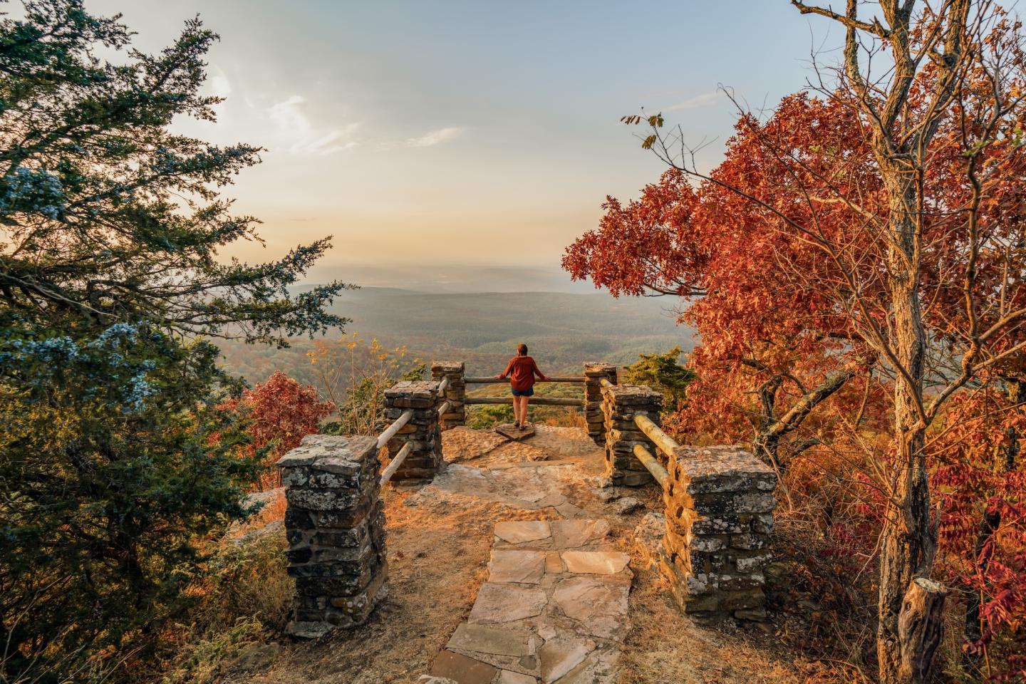 Person stands on scenic overlook, surrounded by autumn trees and distant mountains.