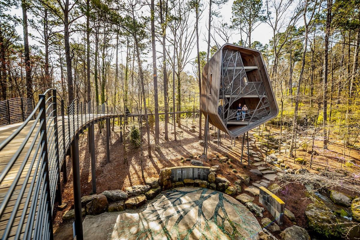 A modern treehouse with a pathway in a sunlit forest setting.