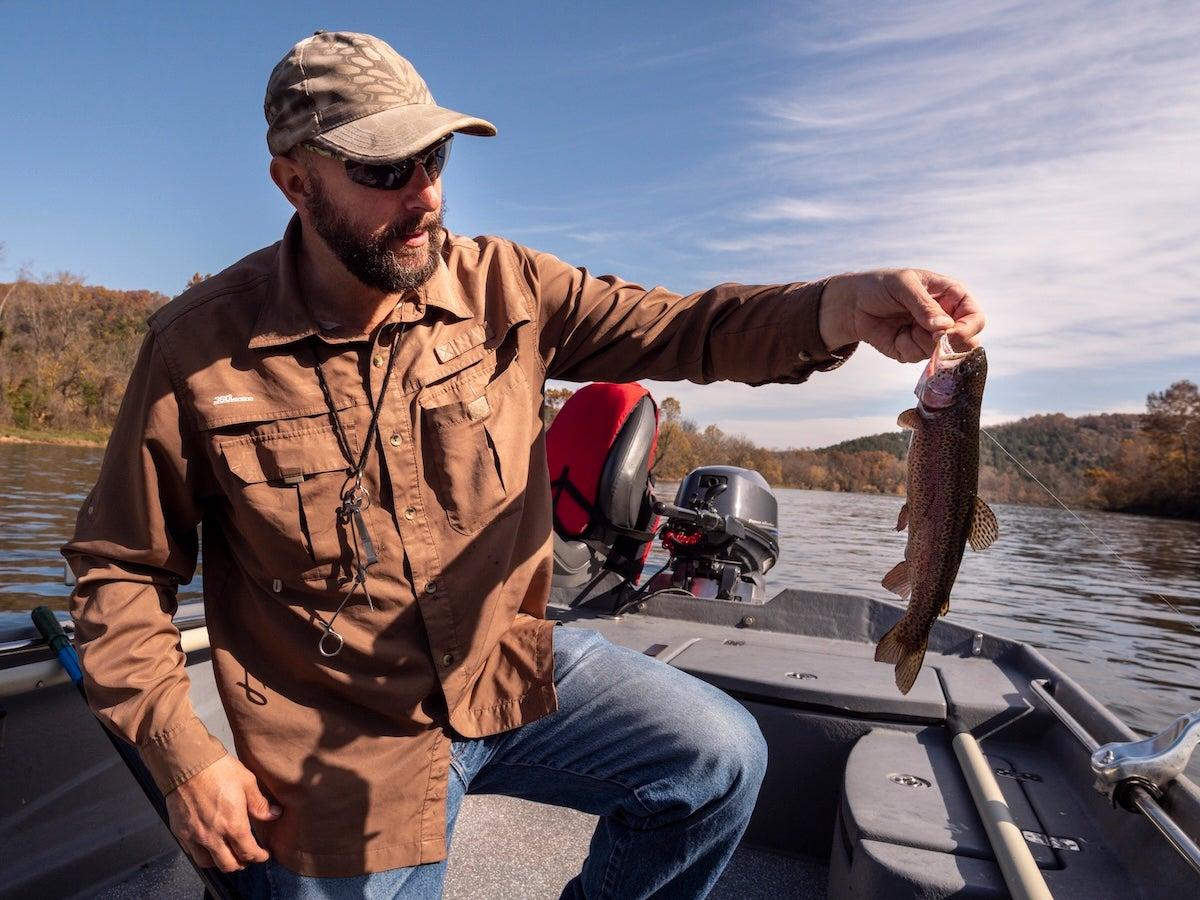 Man in a boat holds a fish, wearing a brown jacket and cap, with lake and trees in background.
