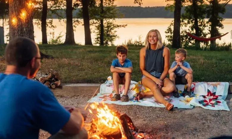 Family gathered around a campfire by a lake at sunset, with trees in the background.