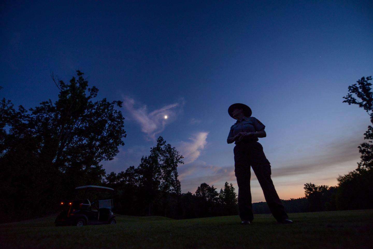 Silhouette of a person at dusk, with a golf cart and moon in the sky.