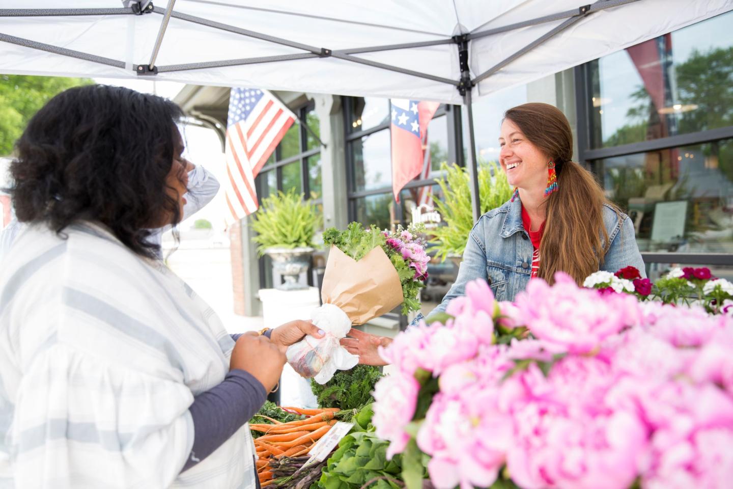 Two women smiling at a market stall, surrounded by flowers and flags.