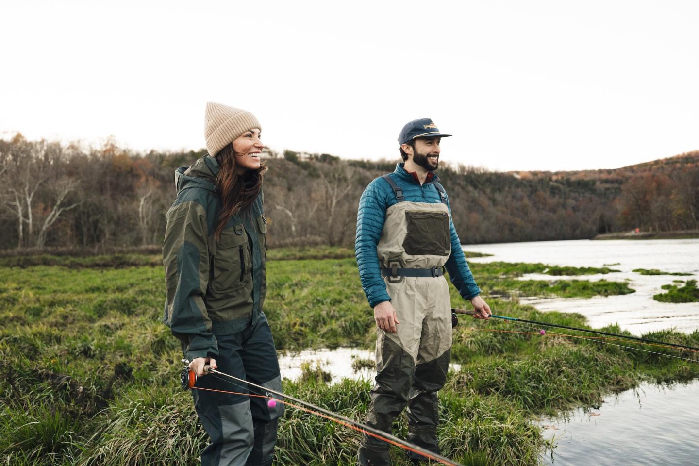 Two people fishing by a river, wearing outdoor gear and smiling.