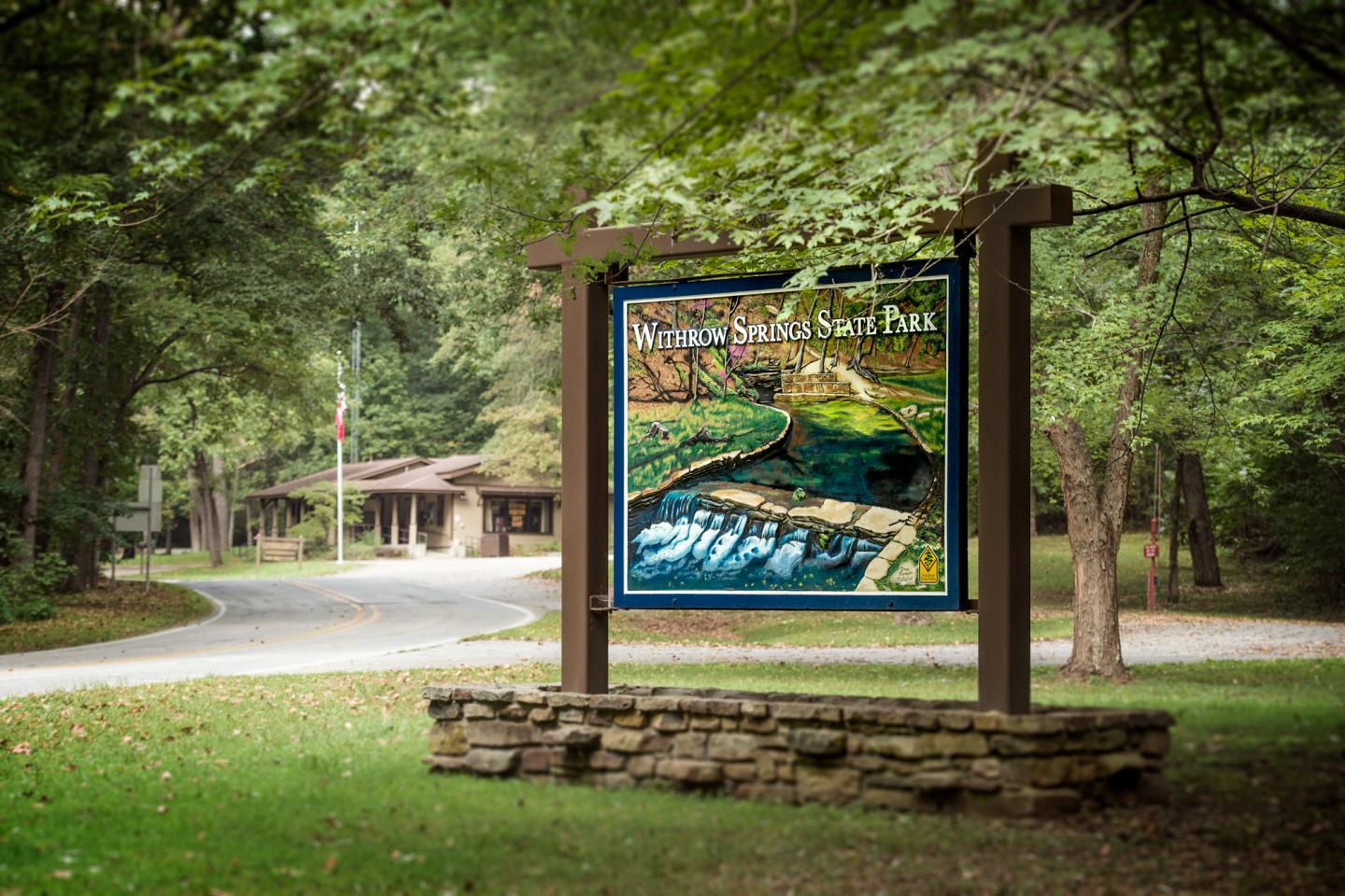 Park entrance sign with colorful map, surrounded by trees and a road.
