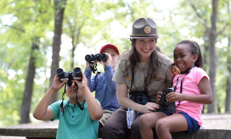 Park ranger with children using binoculars in a forest setting.