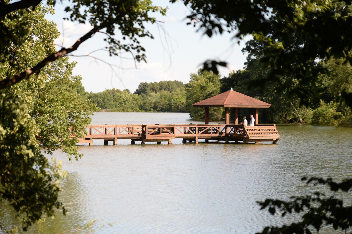 Gazebo on a wooden pier surrounded by trees over a serene lake.