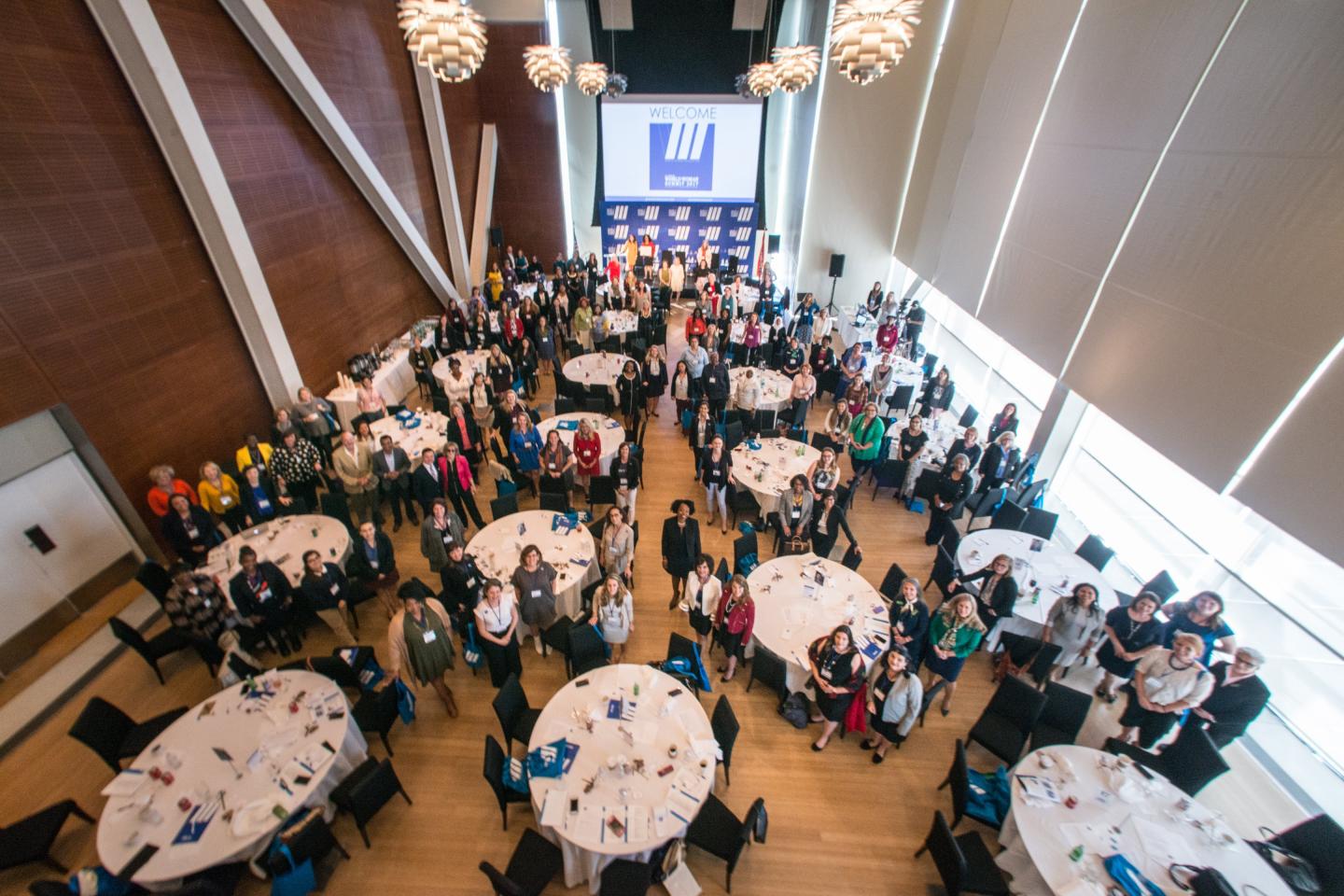 Large group gathered in a conference hall with round tables and a stage at the front.