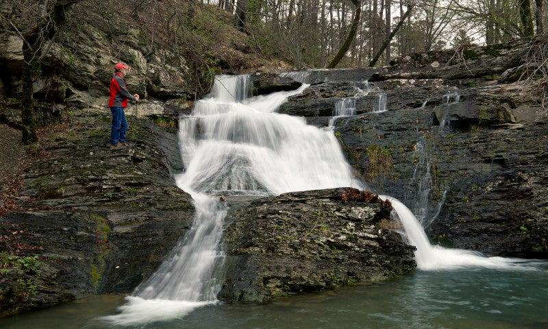Waterfall cascading over rocks in a forest, person in red jacket standing nearby.