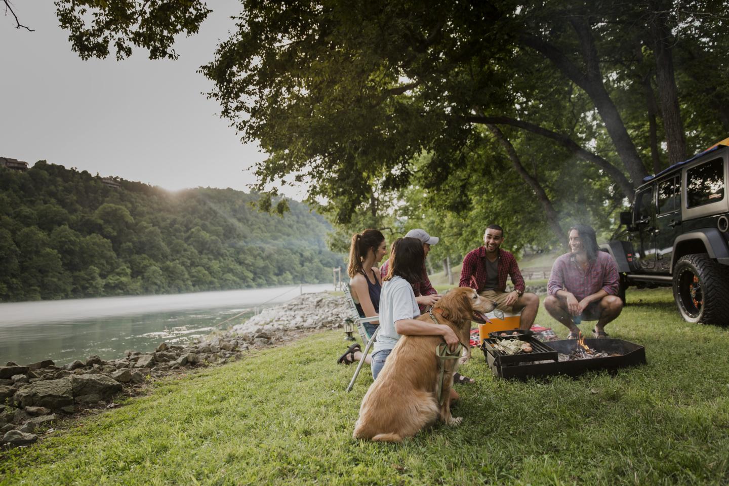 Four friends relax around a campfire with a dog by a river in the countryside at sunset.