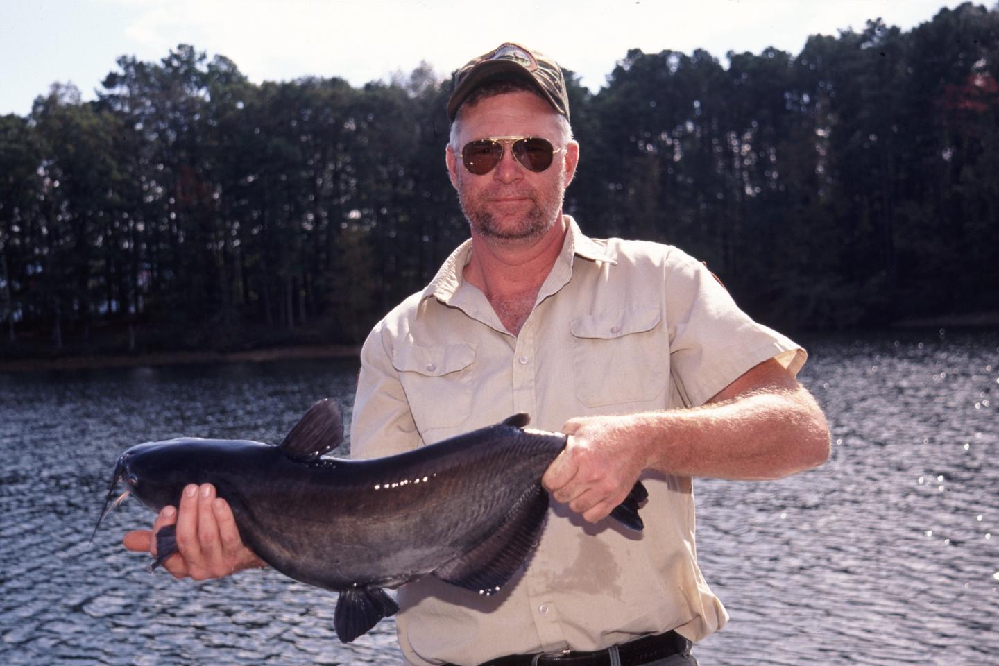 Man holding a large catfish by a lake, wearing sunglasses and a cap.