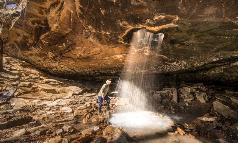 Hiker standing under a waterfall in a rocky cave.
