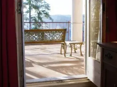 Open door leading to a sunlit balcony with a bench and table. Forested view in the distance.