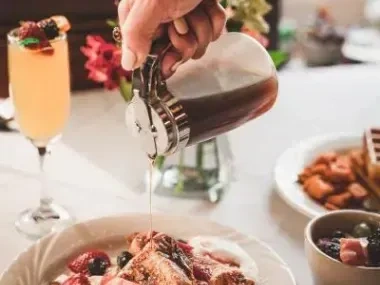 Pouring syrup on waffles with fruit, mimosa in background.