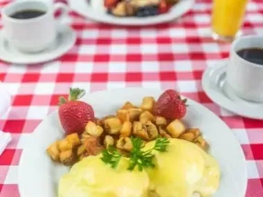Eggs Benedict, potatoes, and strawberries on a plate on a red checkered tablecloth.