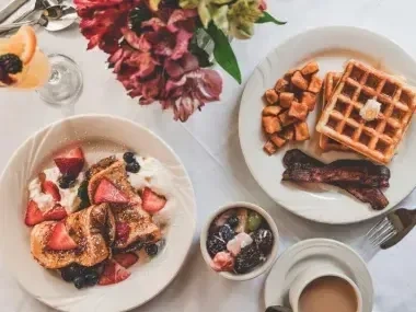 Waffles, pancakes with berries, bacon, and coffee on a table with flowers.