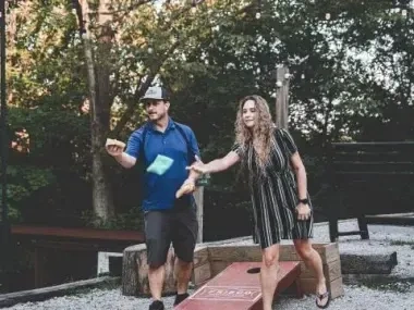 Man and woman playing cornhole outdoors, trees in the background.