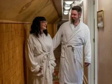 A man and woman in bathrobes smile in a cozy hallway.