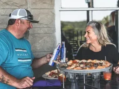 A man and woman smiling over a large pizza on a patio.