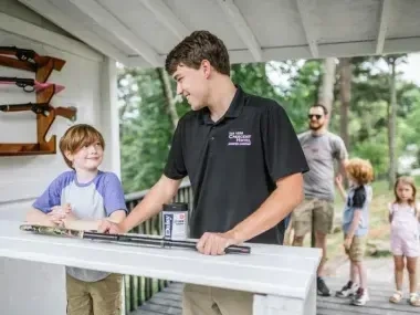 Young boy and man at a shooting range counter, with guns on display, outdoor setting.