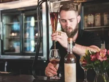 Bartender pouring drink into glass, with flowers and a bottle on the counter.