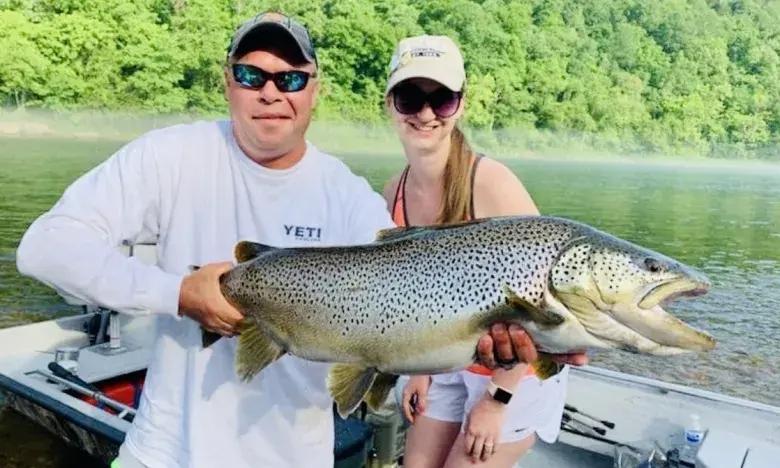 Man and woman on boat holding a large fish, with green trees in the background.