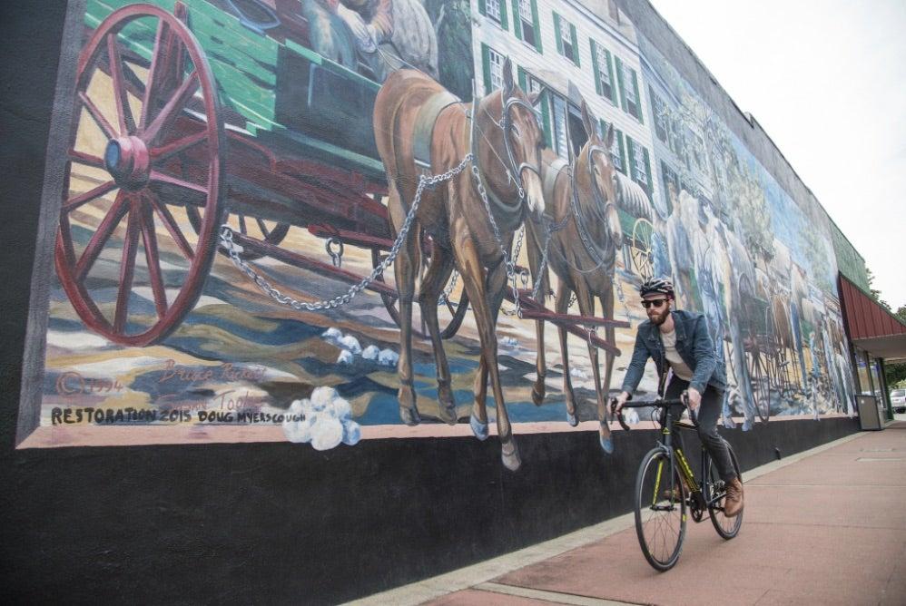 Cyclist rides past large mural of historical horse-drawn carriage.