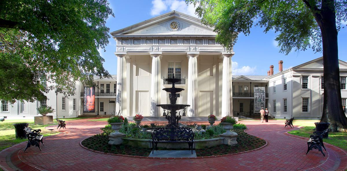 Grand building with columns, fountain in courtyard, and benches under trees.