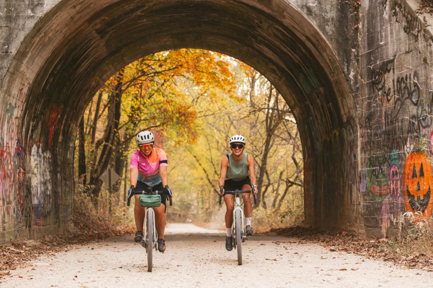 Cyclists ride under a graffiti-covered bridge with autumn trees in the background.
