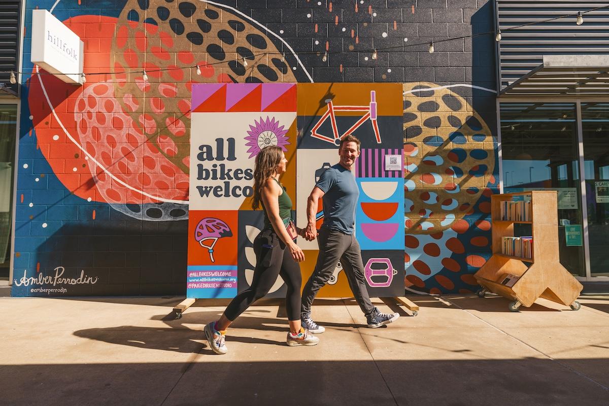 Two people walk past a colorful mural in bright sunlight.