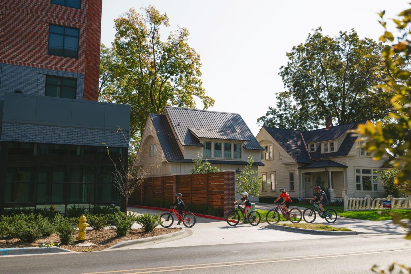 Cyclists ride past houses on a sunny suburban street.