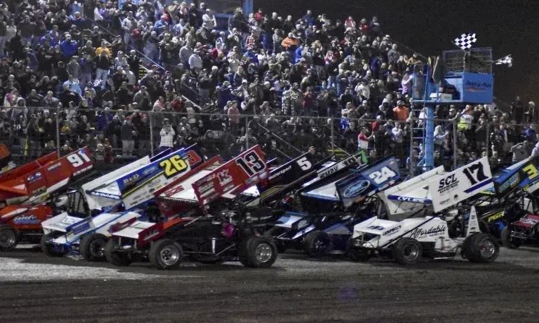 Race cars lined up on a dirt track with a large crowd in the stands.