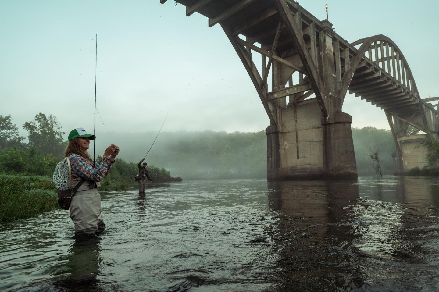 Under a bridge, people wade in a river, fishing with rods.