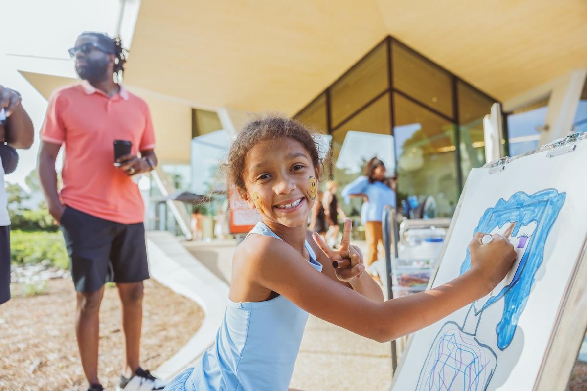 Child drawing on easel outside, adults nearby under modern canopy.