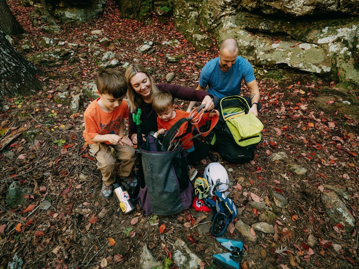 Family sits in a forest with backpacks and climbing gear on the ground.