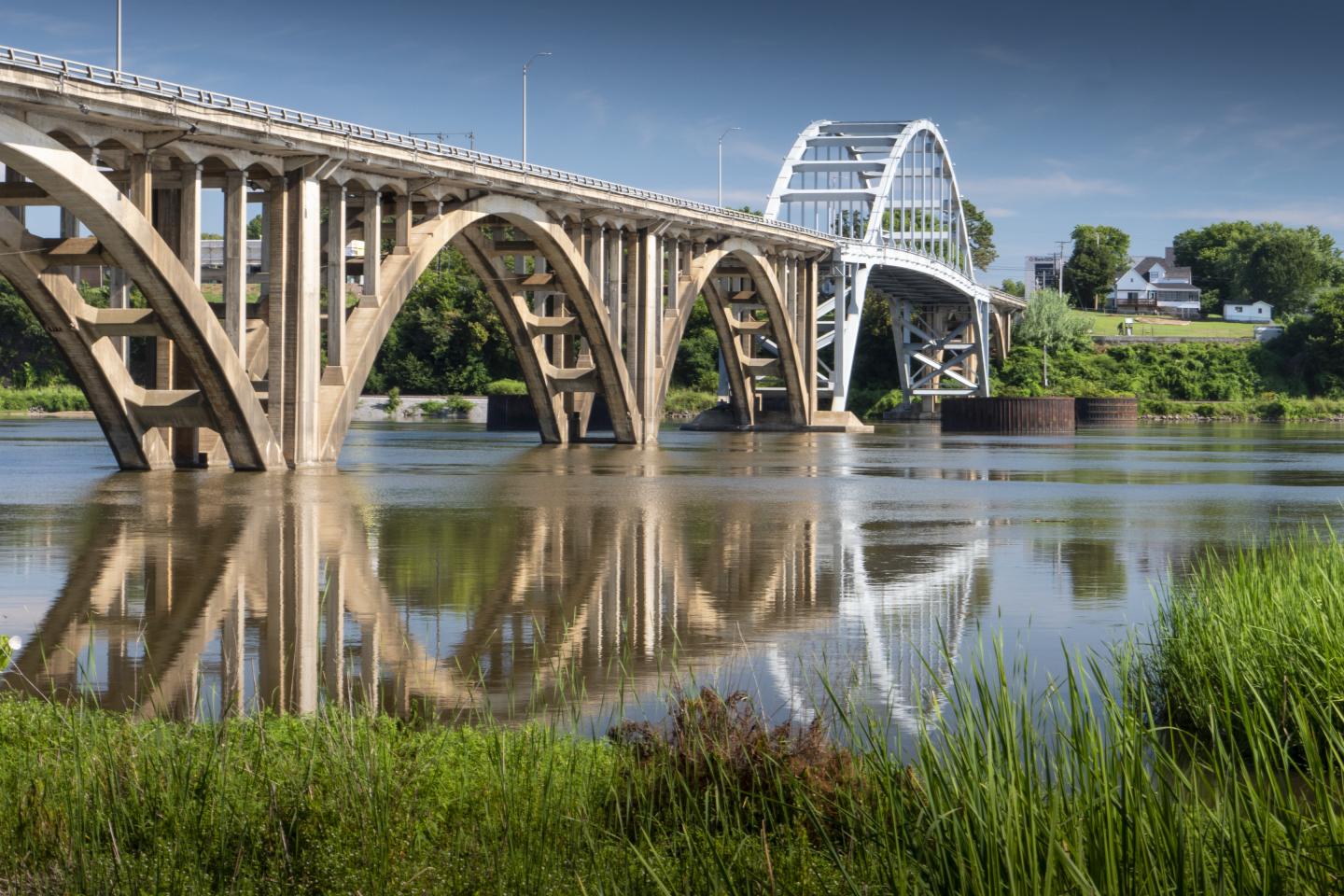 Arched bridge reflected in calm river, surrounded by greenery.