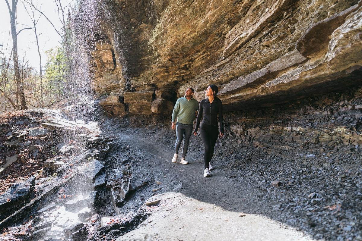 Two people walking on a rocky trail under a cliff, with sunlight filtering through trees.