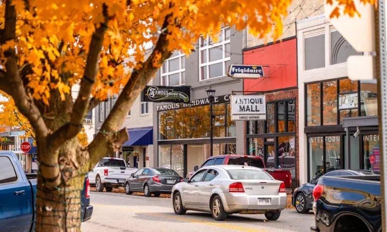 Street with fall foliage, parked cars, and storefronts.
