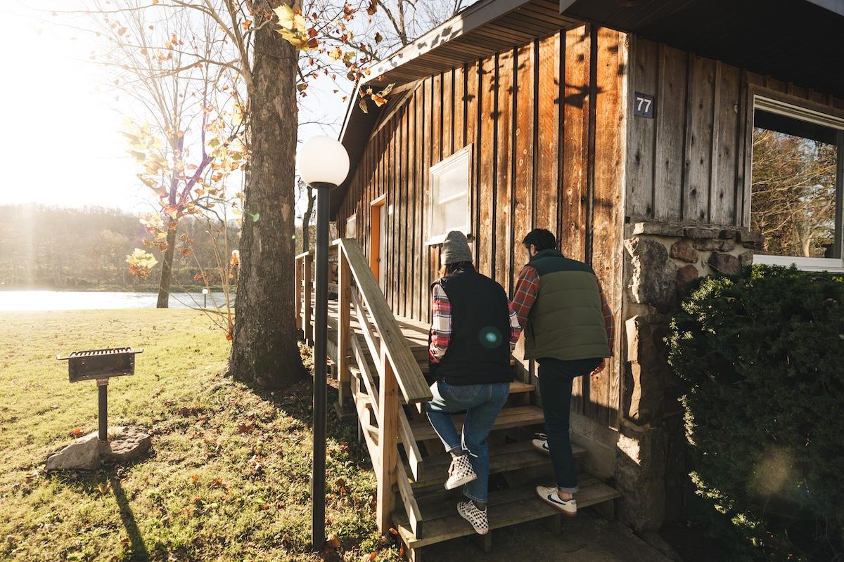 Two people walking up steps to a rustic wooden cabin in sunlight.