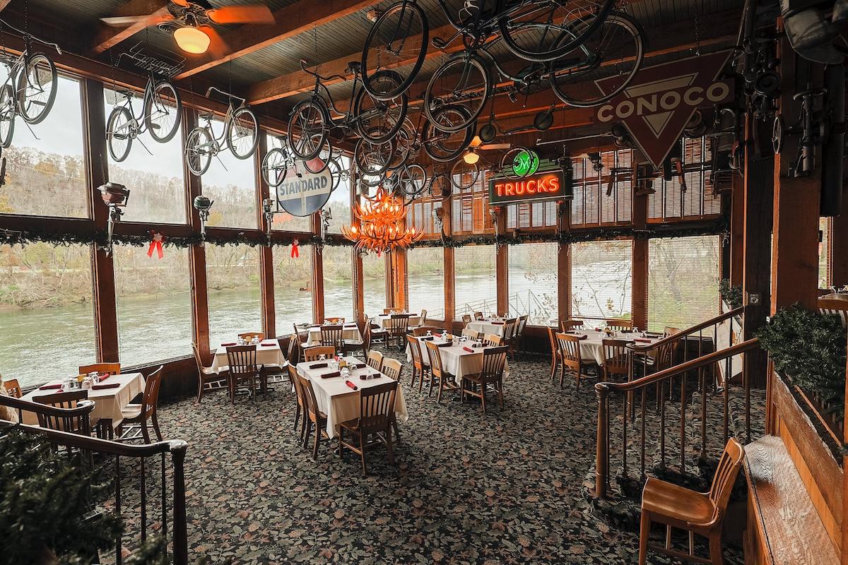 Cozy restaurant interior with bicycles hanging from ceiling, large windows, and river view.