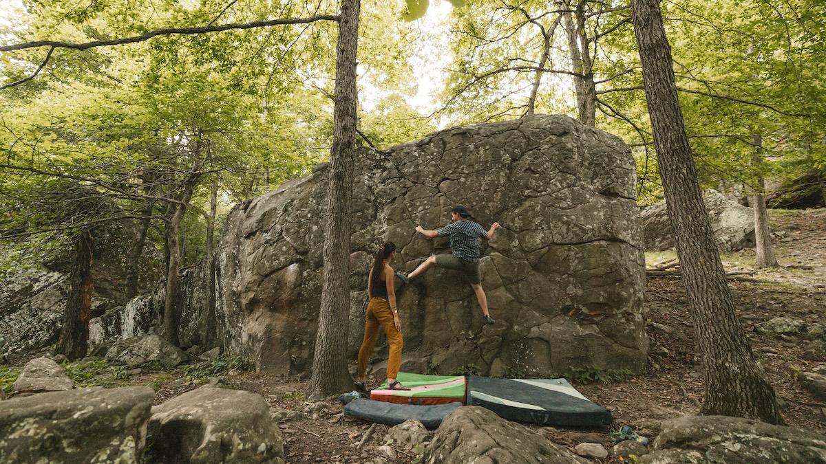 Two people bouldering on a rocky wall in a forest setting with climbing mats below.