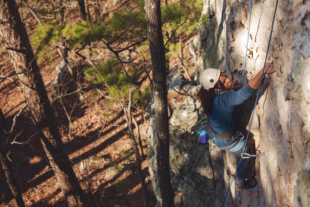 Rock climber scaling a tall cliff, wearing a helmet, surrounded by trees.